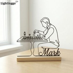 May include: A black metal chess-themed sculpture depicting a person playing chess. The sculpture is mounted on a wooden base with the name "Mark" in cursive. The chess pieces and the person are rendered in a continuous line style.