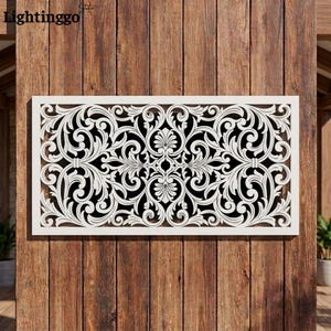 May include: A rectangular white wall panel featuring an intricate, symmetrical laser-cut scrollwork pattern with swirling acanthus leaf motifs. The design is framed by a solid white border and is mounted against a vertical brown wood plank wall.