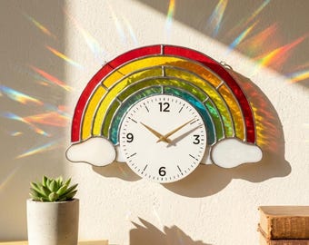 May include: A rainbow-shaped wall clock with a white clock face and gold hands. The clock features a colourful stained-glass design in red, yellow, green, and blue. The clock is mounted on a white wall.