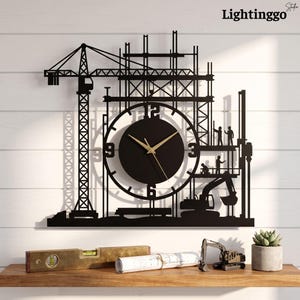 May include: A black wall clock featuring a construction site silhouette with a crane, scaffolding, and workers. The circular clock face has gold-colored hands and large numerals at the 3, 6, 9, and 12 positions, integrated into the metal structure.