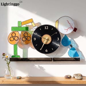 May include: A wall clock featuring a central black vinyl record face with white numerals, flanked by stained glass elements depicting an optometry phoropter on the left and a human eye with blue teardrop shapes on the right, all on a dark base.
