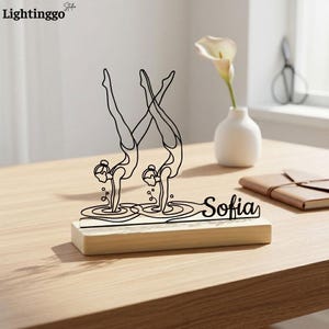 May include: A wooden desk ornament featuring two stylized gymnasts in a handstand pose. The black line art design includes the name "Sofia" and is set on a light-colored wooden base. The "Lightinggo" logo is in the top left corner.