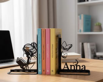 Puede incluir: Un par de sujetalibros de metal negro con la silueta de una persona leyendo un libro a la izquierda y el nombre Anna con dos iconos de libros abiertos a la derecha, sosteniendo cuatro libros coloridos en posición vertical sobre madera.