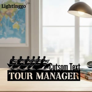 May include: A black wooden sign featuring a silhouette of a crew team rowing in a boat. The text "TOUR MANAGER" is displayed below the boat, with the words "Cutsom Text" above the boat. The sign is likely a decorative item.