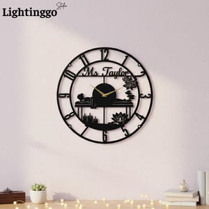 May include: Black metal wall clock with a spa theme. The clock features Roman numerals, a spa scene with a person, candles, and lotus flowers. The clock hands are gold, and the name "Ms. Taylor" is above the clock face.