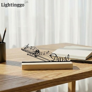 Puede incluir: Un trombón de metal negro con notas musicales y el nombre "David" sobre una base de madera. La pieza decorativa está sobre un escritorio de madera con un lápiz y un cuaderno en el fondo.