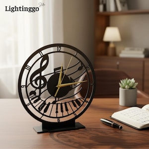 May include: Black metal desk clock with a musical theme. The clock features a treble clef, musical notes, and piano keys within a circular frame. Gold-coloured hands indicate the time. The clock sits on a black rectangular base.