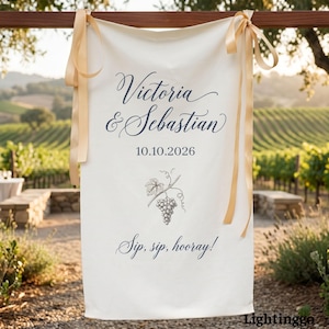 May include: Cream-colored wedding banner with the names "Victoria & Sebastian" and the date "10.10.2026" in navy blue script. A grape vine illustration is below the date, with the phrase "Sip, sip, hooray!" at the bottom. Gold ribbons are tied at the top.