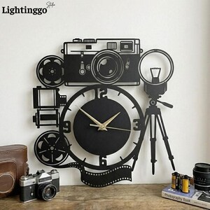 Puede incluir: Reloj de pared de metal negro con forma de cámara vintage, con una esfera en el centro. El reloj presenta una cámara, carretes de película y un trípode. Las manecillas del reloj son doradas. La palabra "Lightinggo" está en la esquina superior izquierda.