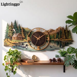 May include: A wooden wall clock featuring a layered landscape scene with a log cabin, pine trees, mountains, and a lake with two loons. The clock face is circular with gold-colored numbers and hands, set against a dark brown background.