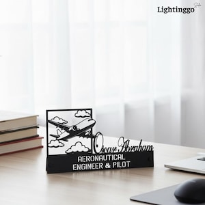 Può includere: Organizzatore da scrivania in metallo nero con un design traforato di un aereo che vola tra le nuvole. L'organizzatore include il nome "Oscar Abraham" e il testo "AERONAUTICAL ENGINEER & PILOT". Il logo "Lightinggo" è visibile in alto a destra.