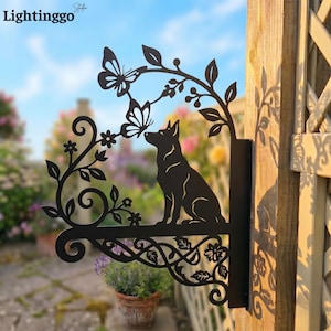Op de afbeelding: Zwarte metalen tuinversiering met een hondensilhouet, vlinders en bloemmotieven. Het decoratieve stuk toont een hond die naar een vlinder kijkt, met een decoratief bloemen- en bladerontwerp. Het bord is op een houten paal gemonteerd.