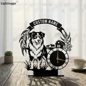 May include: Black laser-cut decorative clock featuring a border collie dog, a barn, and the words "CUSTOM NAME". The clock has a round face with Roman numerals and gold-colored hands. The clock is on a wooden surface.