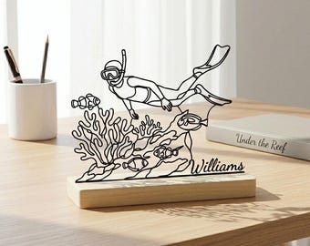 May include: Black metal sculpture depicting a scuba diver, coral, and fish on a wooden base. The diver wears a mask and fins. The base is engraved with the name "Williams". A decorative item for the home.