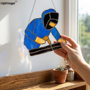 May include: Stained glass art piece depicting a welder in blue, black, and yellow. The artisan is shown working on a black surface. The piece is suspended by a silver chain. The image is set against a white wall and a window.