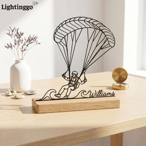 May include: A black metal sculpture of a paraglider with a parachute above a wave design, mounted on a wooden base. The name "Williams" is written on the base. The sculpture is on a light-coloured wooden table.