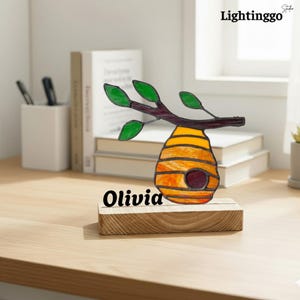 Op de afbeelding: Een gebrandschilderde bijenkorf op een houten voet, met een bruine tak en groene bladeren. De bijenkorf is geel en oranje met een donkerrode opening. De naam "Olivia" staat op de voet.