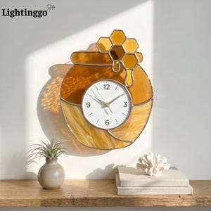 May include: A stained glass wall clock shaped like a honey pot with a honeycomb design. The clock face is white with black numbers and gold hands. The clock is mounted on a white wall.