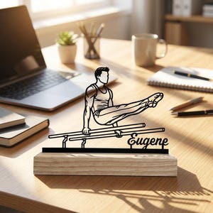 May include: A black metal silhouette of a gymnast performing on parallel bars, mounted on a rectangular light wood base. The figure is depicted in a horizontal position, and the name Eugene is written in a cursive script on the base structure.