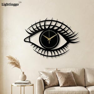Puede incluir: Reloj de pared negro con forma de ojo, con esfera circular y manecillas doradas. El reloj tiene un diseño decorativo de pestañas. La marca "Lightinggo" está en la esquina superior izquierda.