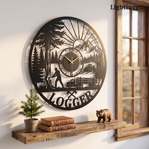 May include: A circular black metal wall clock featuring a laser-cut design of a logger felling a tree, a logging truck, mountains, and pine trees. The word LOGGER is cut into the bottom with two crossed axes. It has gold-colored clock hands.