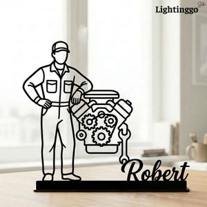 May include: Black metal silhouette of a mechanic with an engine and the name "Robert". The mechanic is wearing a cap and overalls. The engine features gears and a wrench. The product is a decorative item.