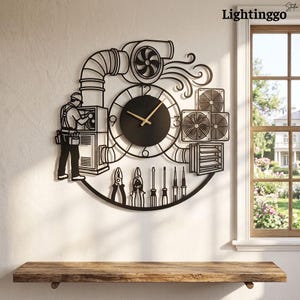 Könnte beinhalten: Eine Wanduhr aus schwarzem Metall mit einem zentralen runden Zifferblatt und goldenen Zeigern. Das Design zeigt die Silhouette eines Technikers an einer HLK-Anlage, Lüftungskanäle, einen Ventilator sowie verschiedene Werkzeuge im unteren Bereich.