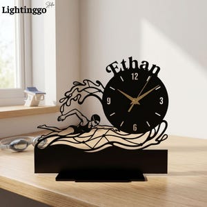 May include: A black tabletop clock featuring a silhouette of a swimmer in motion amidst stylized waves. The clock face is circular with gold-colored hands and the name Ethan displayed in a decorative font above the dial on a flat base.