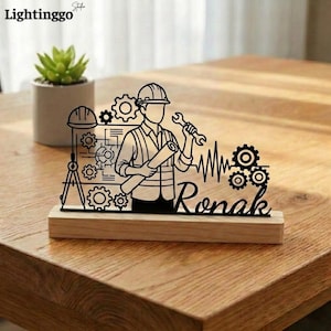 May include: A black laser-cut engineer desk ornament on a wooden base. The design features an engineer holding a wrench and blueprints, with gears, a hard hat, and the name "Ronak". A small succulent plant in a white pot is in the background.