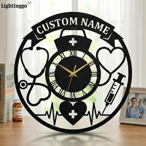 May include: Black metal wall clock with a medical theme. The clock features a nurse's hat, stethoscope, syringe, and heart symbols. The clock face has Roman numerals and gold-colored hands. The text "CUSTOM NAME" is at the top.