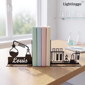 Op de afbeelding: Een paar boekensteunen van zwart metaal met wetenschappelijke laboratoriummotieven. Het linkeronderdeel toont een destilleerapparaat met de naam Louis, terwijl het rechteronderdeel een reageerbuisrek, een Bunsenbrander en een kolf weergeeft.