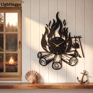 May include: A black metal wall clock shaped like a campfire with flames, logs, and a hanging kettle. The circular clock face features gold-coloured hands and numbers at the 3, 6, 9, and 12 positions. The design is mounted on a white panelled wall.
