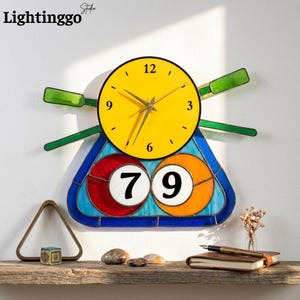 May include: A wall clock featuring a bright yellow circular face with black numerals and hands, positioned above a stained glass panel displaying the numbers 7 and 9 in white circles, flanked by two green glass oars on a light background.