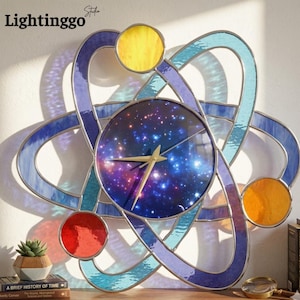 May include: A decorative wall clock with a galaxy-themed face and an atom-like design. The clock features a blue, purple, yellow, and red stained glass design. The clock hands are gold. The text "Lightinggo Studio" is in the upper left corner.