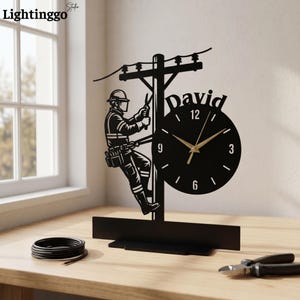 May include: A black metal desk clock featuring a silhouette of a utility worker climbing a power pole. The clock face is circular with white numbers 3, 6, 9, and 12, and gold-colored hands. The name David is cut out above the clock face.