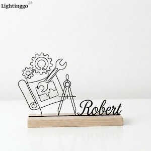 Peut inclure: Une sculpture en fil noir représentant des outils d'ingénierie, notamment des engrenages, une clé et un compas, sur une base en bois. Le nom "Robert" est écrit en script. La sculpture est un objet décoratif.