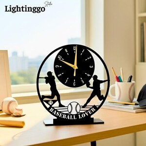 May include: Black baseball-themed clock with gold hands, featuring silhouettes of baseball players and a baseball. The words "BASEBALL LOVER" are below the baseball. The clock is on a black base.