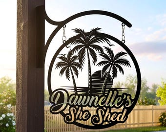 Könnte beinhalten: Schwarzes Metallschild mit einem kreisförmigen Design mit Palmen, einem Sonnenuntergang und dem Text "Dawnelle's She Shed". Das Schild ist an einer dekorativen Halterung aufgehängt und an einem Holzpfosten befestigt. Der Text "Lightinggo" befindet sich in der oberen rechten Ecke.
