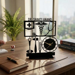 May include: Black laser-cut desk clock with medical symbols and the name "DR. SHAKTI ANAND". The clock features a caduceus, stethoscope, and other medical tools. The clock is on a desk with books and a pen.