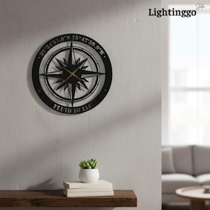 May include: Black metal compass rose wall clock with gold-colored hands. The clock features the text "39°24'57.0"N 75°42'59.9"W" and "FLUID IO LLC" around the outer edge. The clock is mounted on a light gray wall.