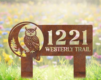 May include: A rusty brown metal address sign displaying the numbers "1221" and the words "Westerly Trail". An owl sits on a branch within a crescent moon and tree design. The sign is in a garden.