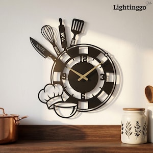 May include: A black metal wall clock featuring a circular face with gold hands and numbers 3, 6, 9, and 12. The clock is decorated with silhouettes of a chef hat, a bowl, and kitchen tools labeled KNIFE, WHISK, ROLL, and SPAT at the top.