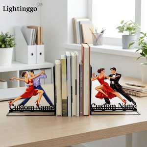 May include: A pair of black metal bookends featuring a cutout illustration of a couple dancing. The figures are depicted in a dynamic pose with the person in the red dress and the partner in a suit. The base includes the text Custom Name.