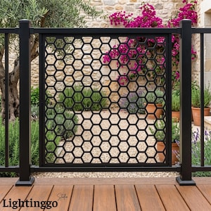 May include: A black metal railing panel featuring a repeating hexagonal honeycomb pattern. The rectangular panel is framed by vertical posts and is positioned above a wooden deck, with a garden and stone wall visible through the open design.