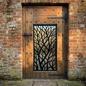 May include: A rectangular wooden door featuring a central vertical panel with a black metal insert displaying an intricate, laser-cut tree branch silhouette pattern. The door is constructed with visible iron hinges and studs on the frame.