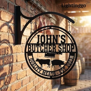 May include: A circular black metal hanging sign featuring the text JOHN'S BUTCHER SHOP, a cleaver icon, silhouettes of a cow and a pig, and the text QUALITY MEATS EST. 2023. The sign is mounted on a black bracket attached to a brick wall.