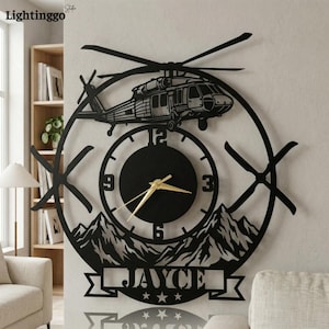 Könnte beinhalten: Schwarze Metall-Wanduhr mit einem Hubschrauber-Design. Das Zifferblatt ist kreisförmig mit goldenen Zeigern, einer Bergkette am unteren Rand und dem Namen "JAYCE" in einem Banner. Die Uhr ist an einer hellen Wand befestigt.