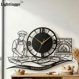 Può includere: Orologio da parete in metallo nero a tema panetteria. L'orologio presenta un fornaio, pane, un forno in mattoni e la scritta "ARTUR'S BAKERY". Le lancette dell'orologio sono dorate. L'orologio è montato su un muro bianco.