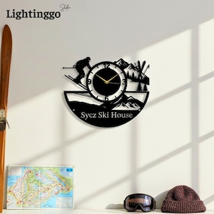 May include: Black metal wall clock featuring a skier, mountains, and ski equipment silhouettes. The clock face has gold hands and a black border. The text "Sycz Ski House" is at the bottom. The brand name "Lightinggo" is at the top left.