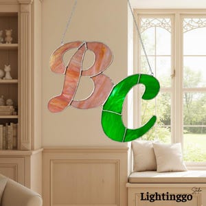 May include: A decorative stained glass piece displaying the letters "BC". The "B" is a mix of pink and orange tones, and the "C" is a bright green. The letters are joined by silver metal and suspended by a chain.